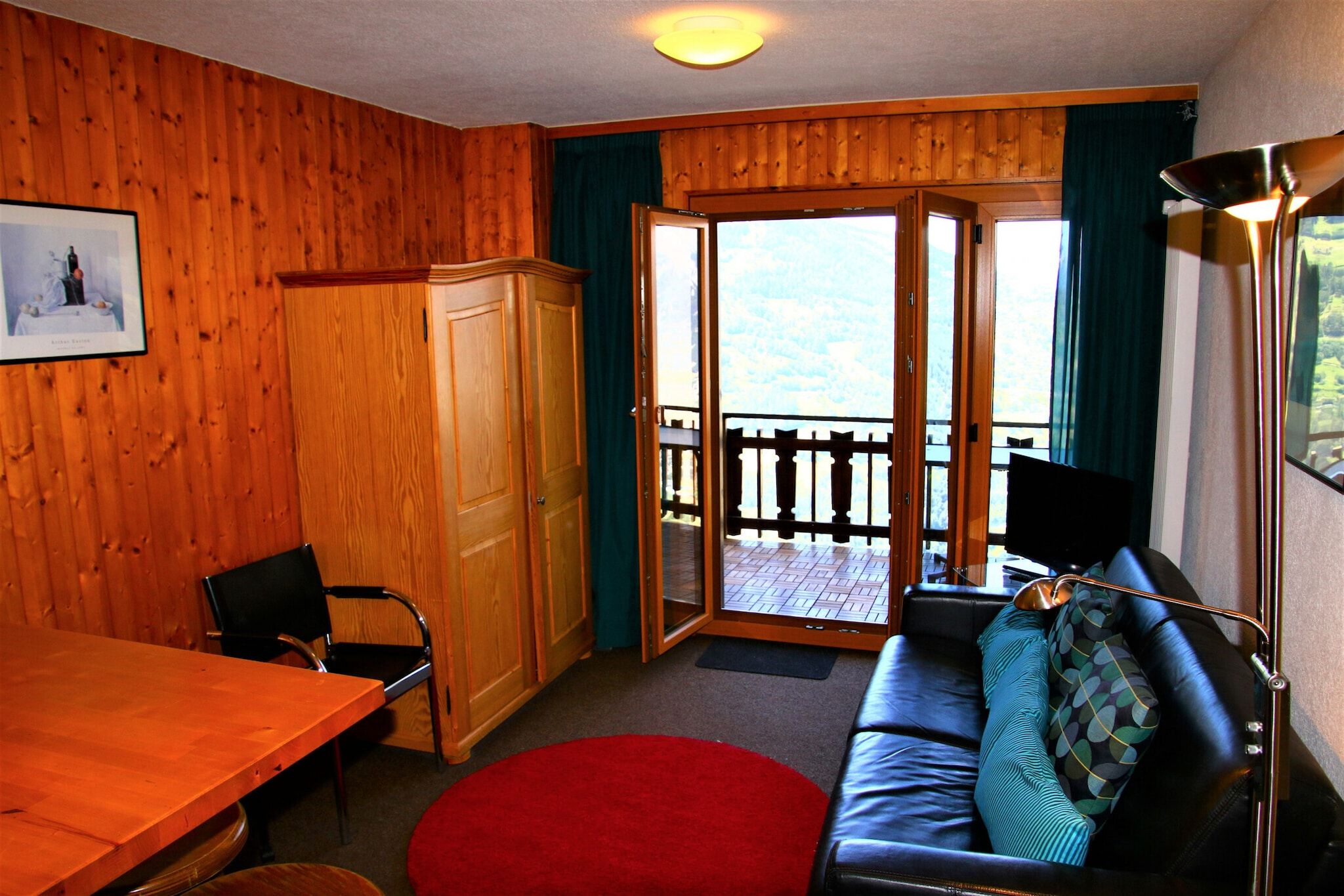 Een kamer met houten details, een eettafel, een bank met kussens, een stoel en een schuifdeur naar een buitenbalkon.