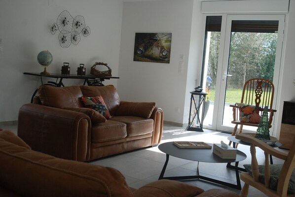 A living room with a brown leather sofa, a small round table, and a wooden chair. There is a globe on a shelf, a painting on the wall, and a window with a view of trees. - La Hal'te Verte, large house on the edge of the voie Verte Sud Ardennes (Rilly-sur-Aisne)