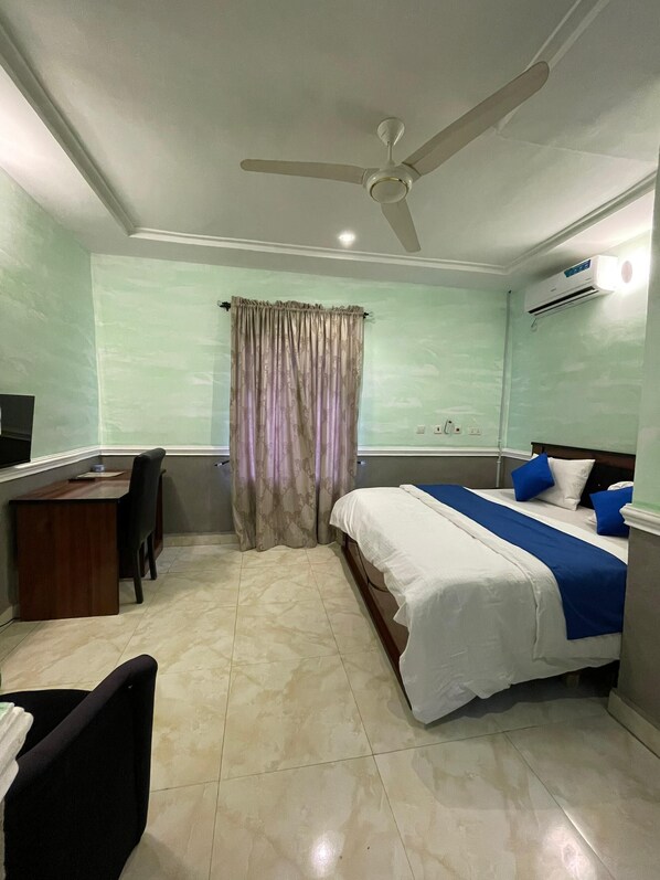 A hotel room with a bed, desk, chair, ceiling fan, and air conditioning unit.