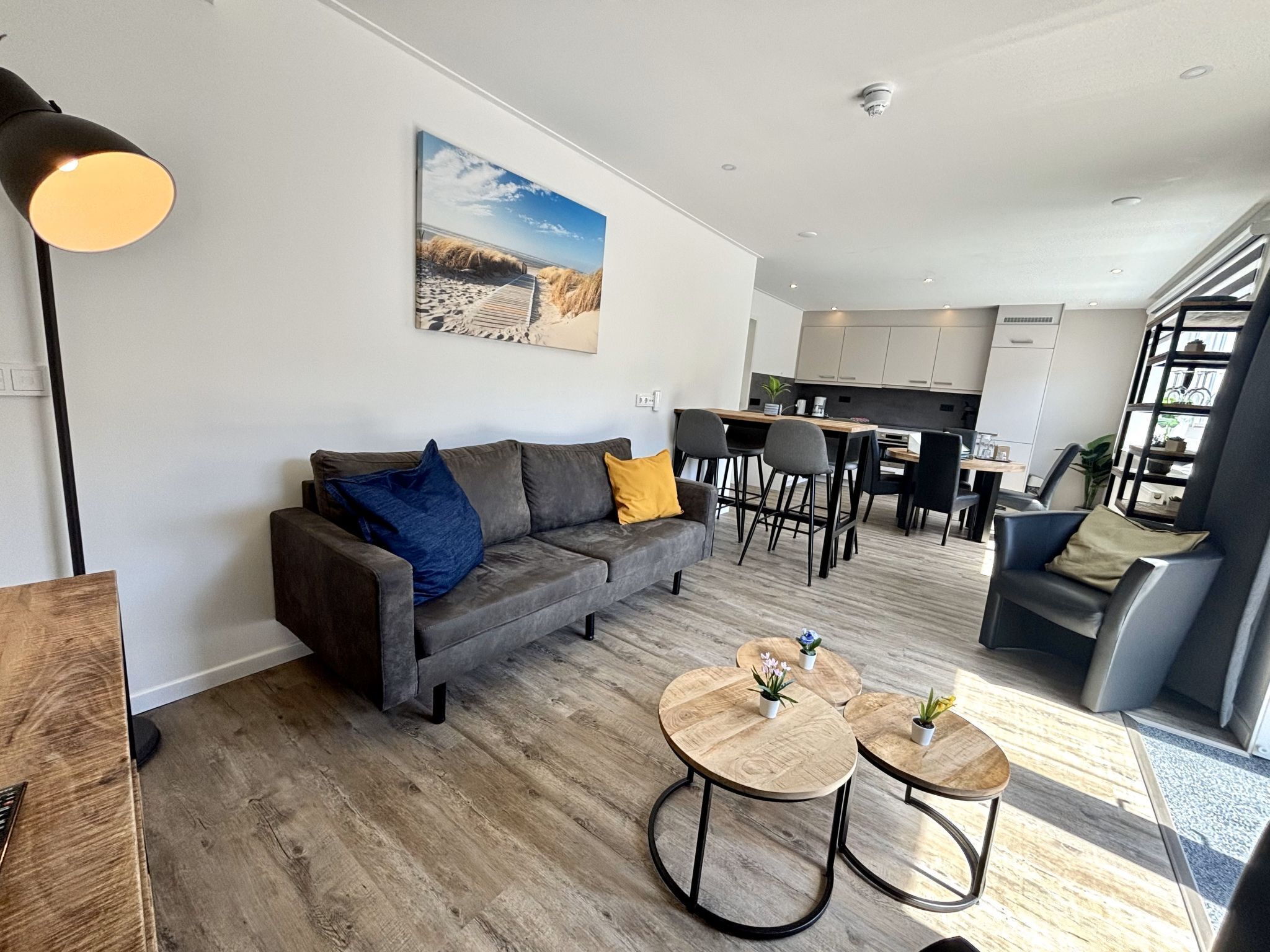 A modern living room with a grey sofa, two round wooden tables, and a dining area with chairs.