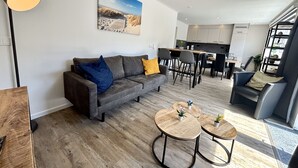 A modern living room with a grey sofa, two round wooden tables, and a dining area with chairs. - Beach House Westkapelle 105.1 by Interhome (Westkapelle)