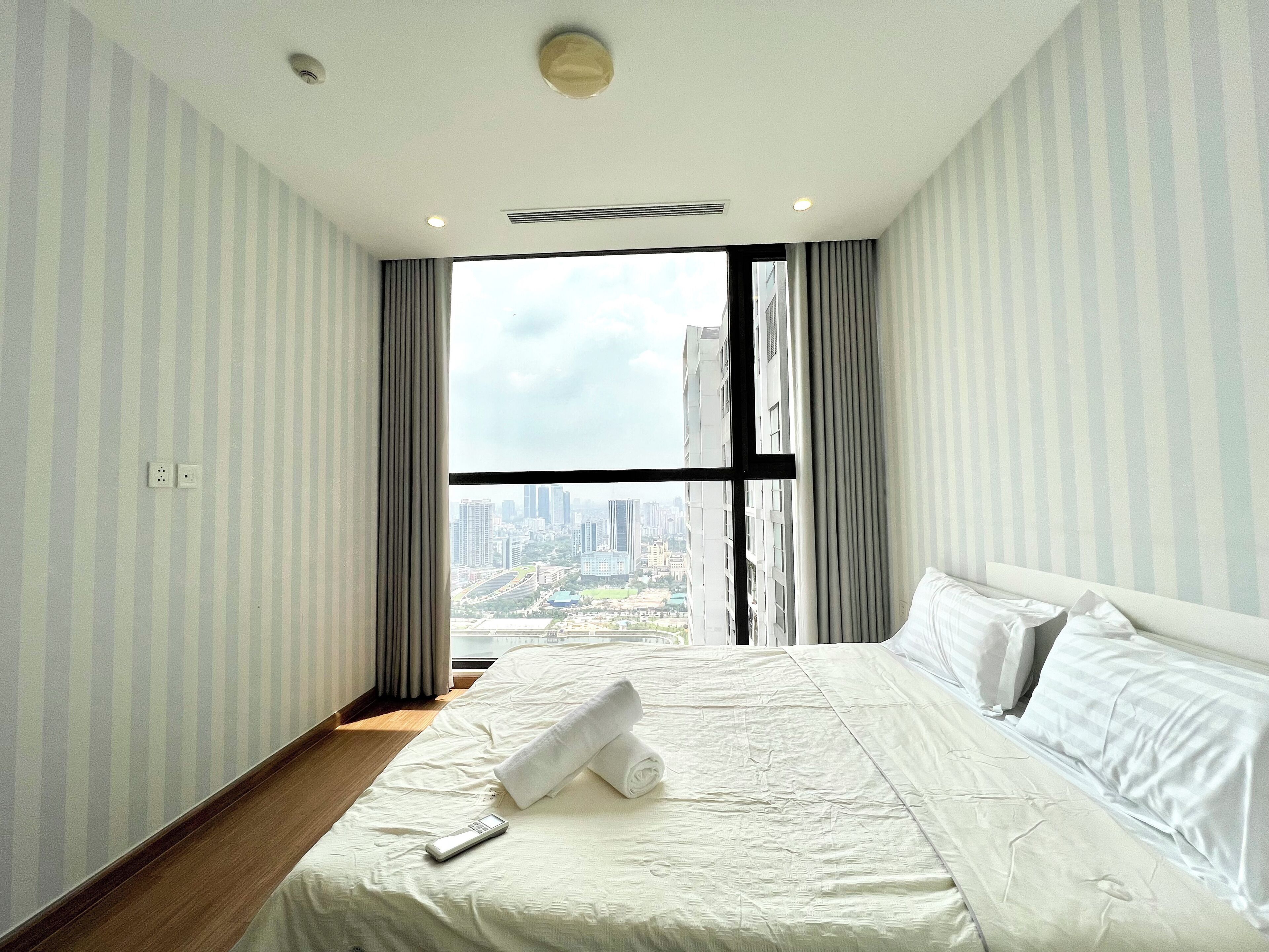Ein Hotelzimmer mit einem großen Bett, einer Fernbedienung und Blick auf eine Stadtlandschaft durch ein großes Fenster.