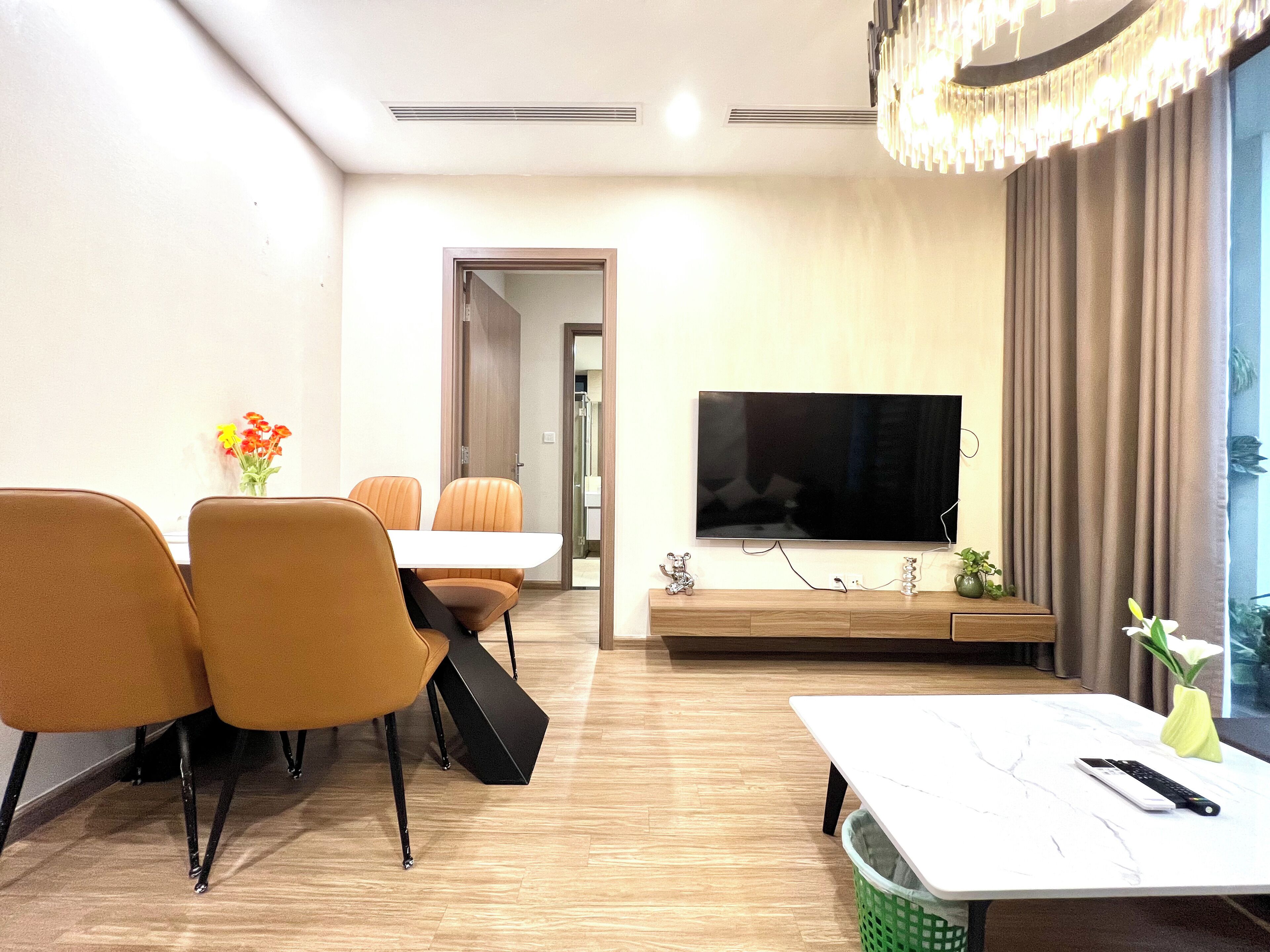A modern living room with a flat-screen TV mounted on the wall, a dining area with orange chairs and a white table, and a small green trash bin on the floor.