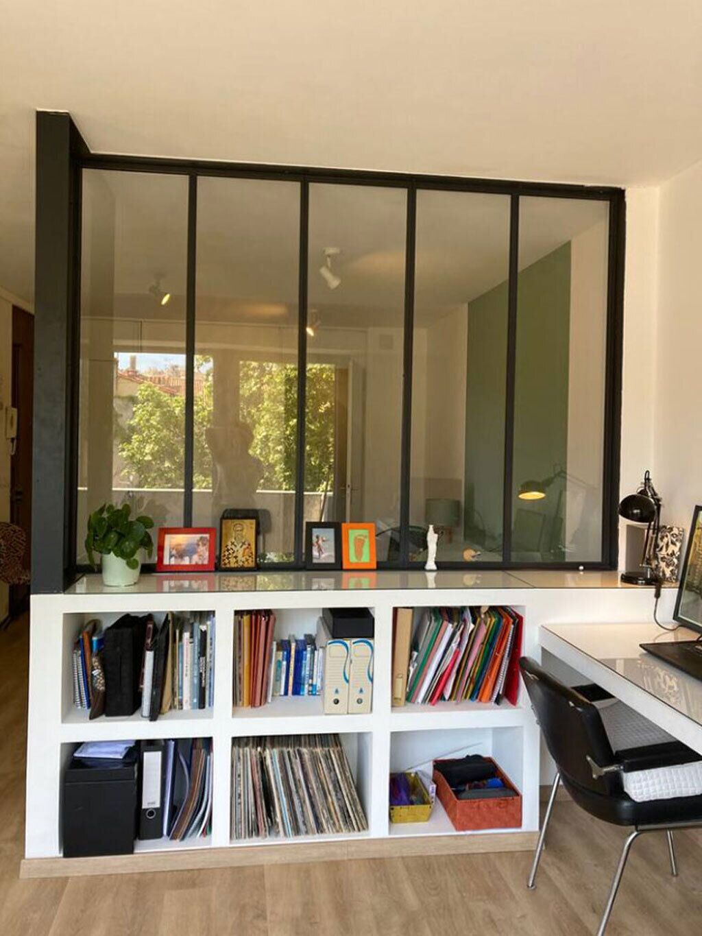 A modern living room with a large glass window, a white bookshelf filled with books and decorative items, a black chair, and a desk with a computer.