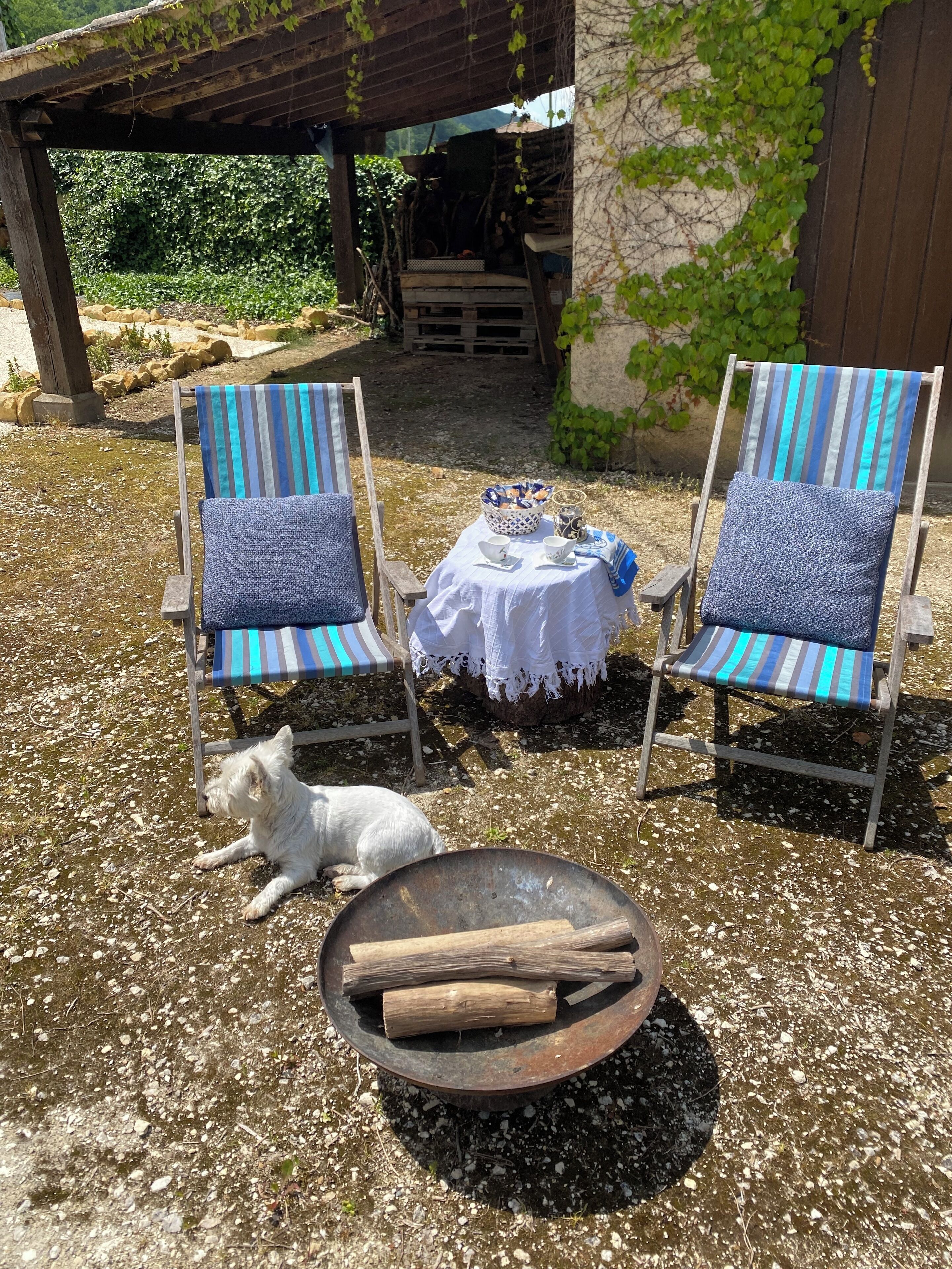 Zwei gestreifte Stühle mit Kissen, ein Tisch mit weißem Tuch und ein Feuerkorb mit Holzscheiten.