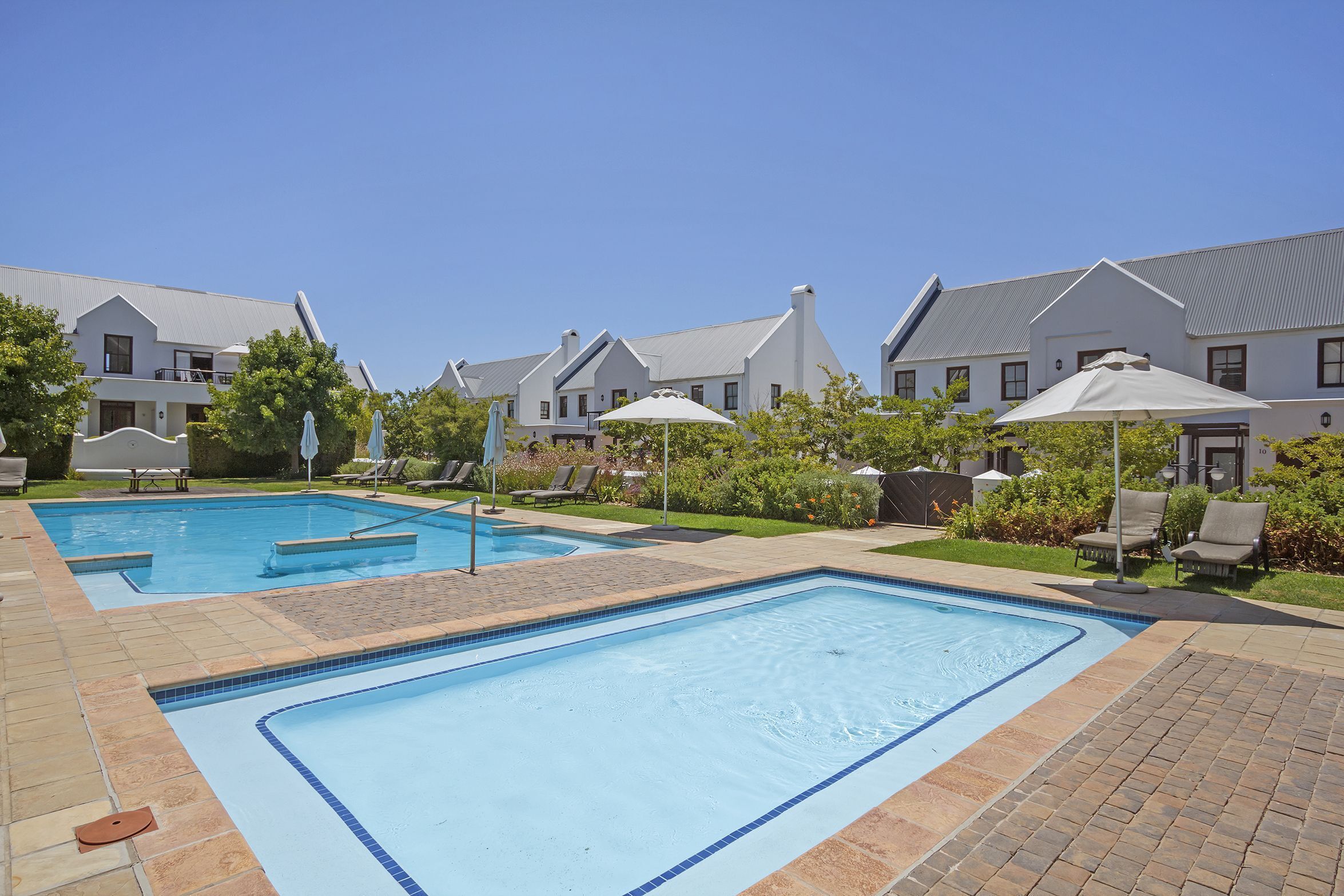 A swimming pool area with two pools, lounge chairs, umbrellas, and a brick pathway.