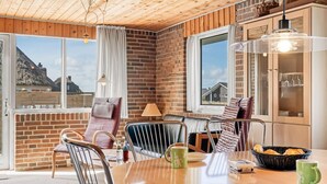 A cozy room with a brick wall, wooden ceiling, and large windows. There is a round table with chairs, a lamp, and a basket of bread. - "Ava" - 350m from the sea by Interhome (Harboøre)
