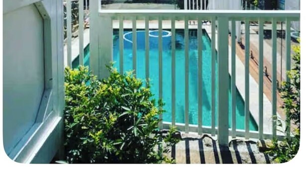 Vista de una piscina desde un balcón con barandilla blanca, rodeado de vegetación y un cielo despejado.