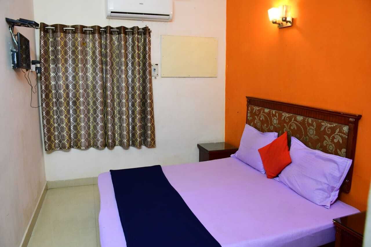 A hotel room with a bed, a nightstand, a wall-mounted air conditioner, a curtain, and a wall-mounted light fixture.