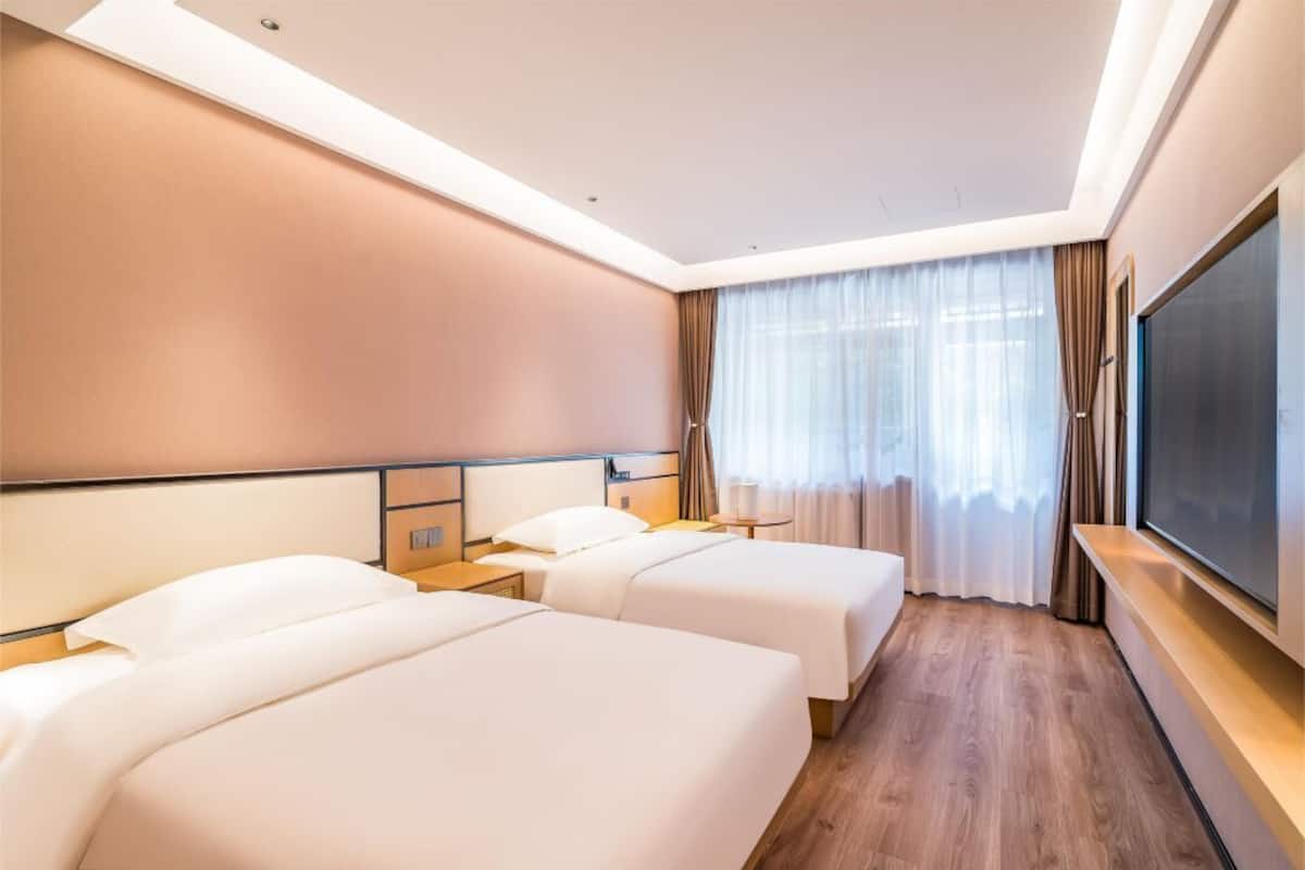 A hotel room with two beds, a large flat-screen TV, a wooden floor, and a large window with sheer curtains.