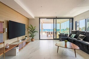 Ein modernes Wohnzimmer mit einem Flachbildfernseher, einer Couch, einem Couchtisch und einer Glastür, die auf einen Balkon mit Meerblick führt.