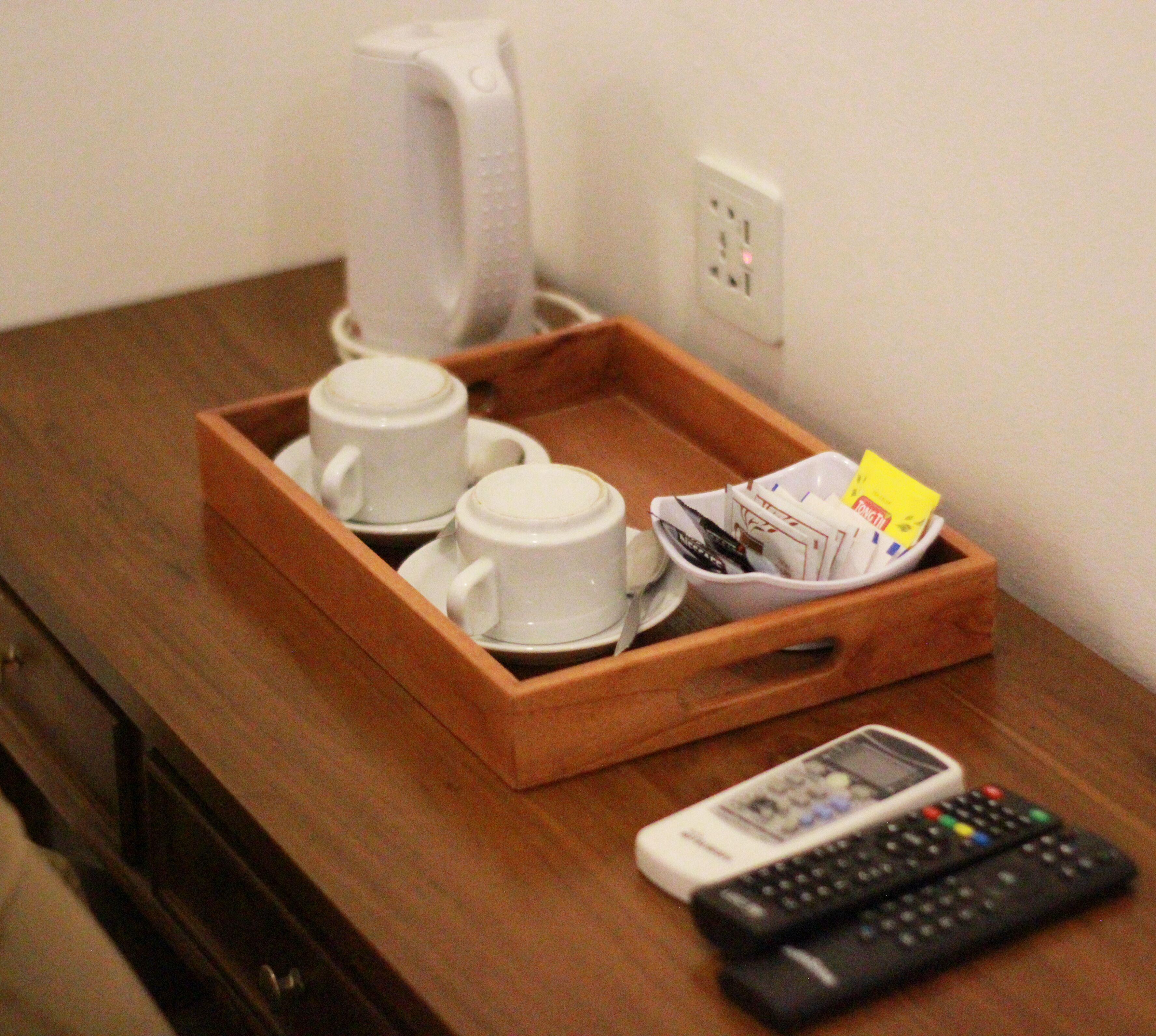 A wooden tray with two white cups and saucers, a coffee maker, a basket of magazines, and two remote controls on a wooden table.