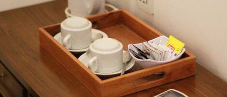 A wooden tray with two white cups and saucers, a coffee maker, a basket of magazines, and two remote controls on a wooden table.