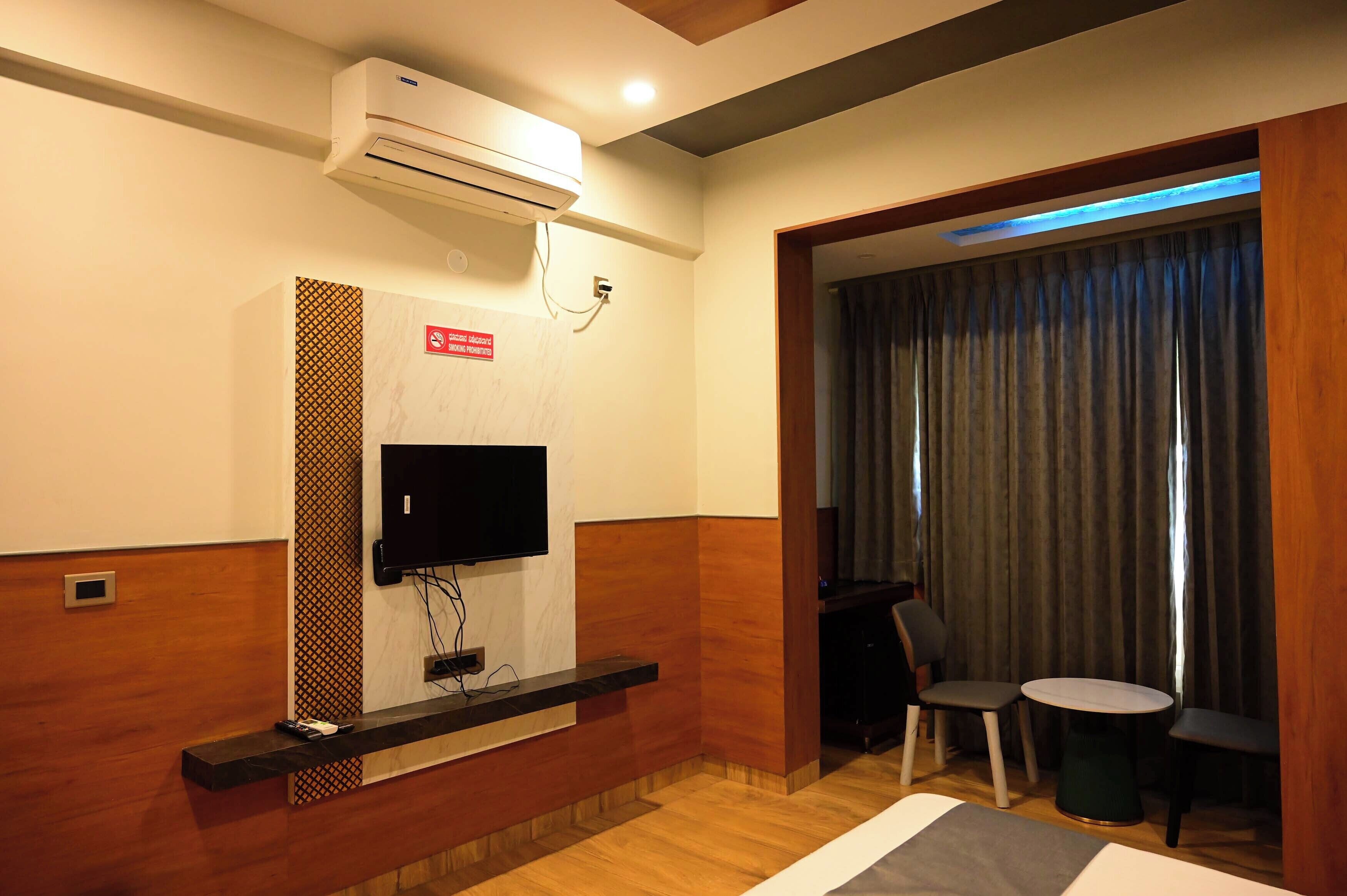A hotel room with a flat-screen TV mounted on a wooden panel, a small table with two chairs, and a large window with curtains.