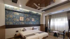 A modern hotel room with a large bed, a ceiling fan, a small table with chairs, and a large window with curtains. - RR Residency (Mysore)
