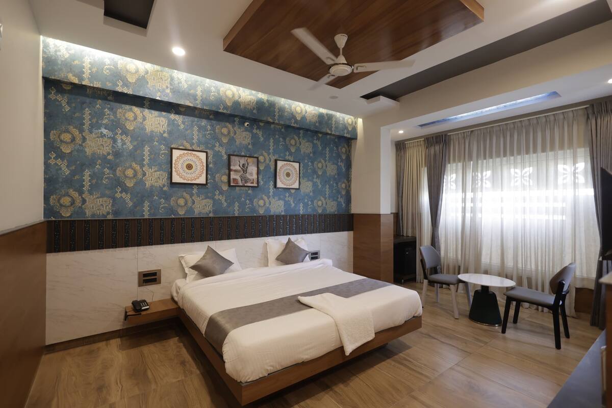 A modern hotel room with a large bed, a ceiling fan, a small table with chairs, and a large window with curtains.