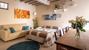 Ein gemütliches Wohnzimmer mit einer Couch, einem Esstisch und einem Couchtisch. An den Wänden hängen zwei Gemälde, eine Uhr und auf dem Esstisch steht eine Vase mit Blumen.