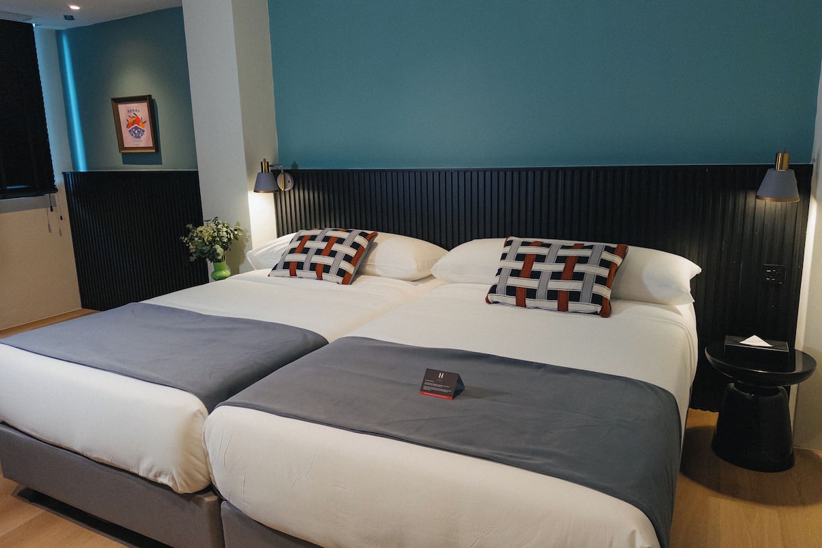 A double bed with two pillows, a bedside table, a lamp, and a small trash bin.
