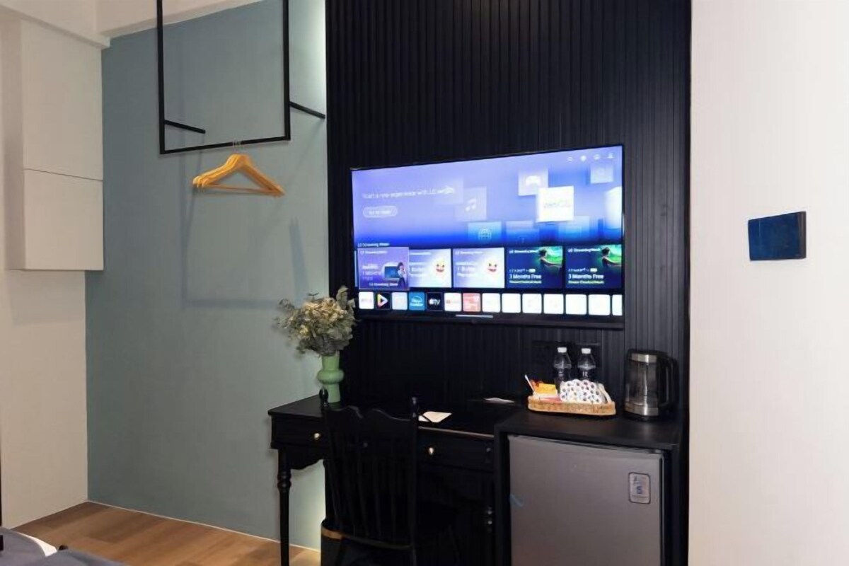 A modern room with a large flat-screen TV mounted on the wall displaying a music streaming service. There is a small desk with a coffee maker, a vase with flowers, and a chair.