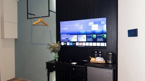 A hotel room with a flat-screen TV mounted on the wall displaying a streaming service interface. There is a small table with a chair, a vase with flowers, and a basket with a coffee maker on it. - Legacy Hill Hotel (Kuching)