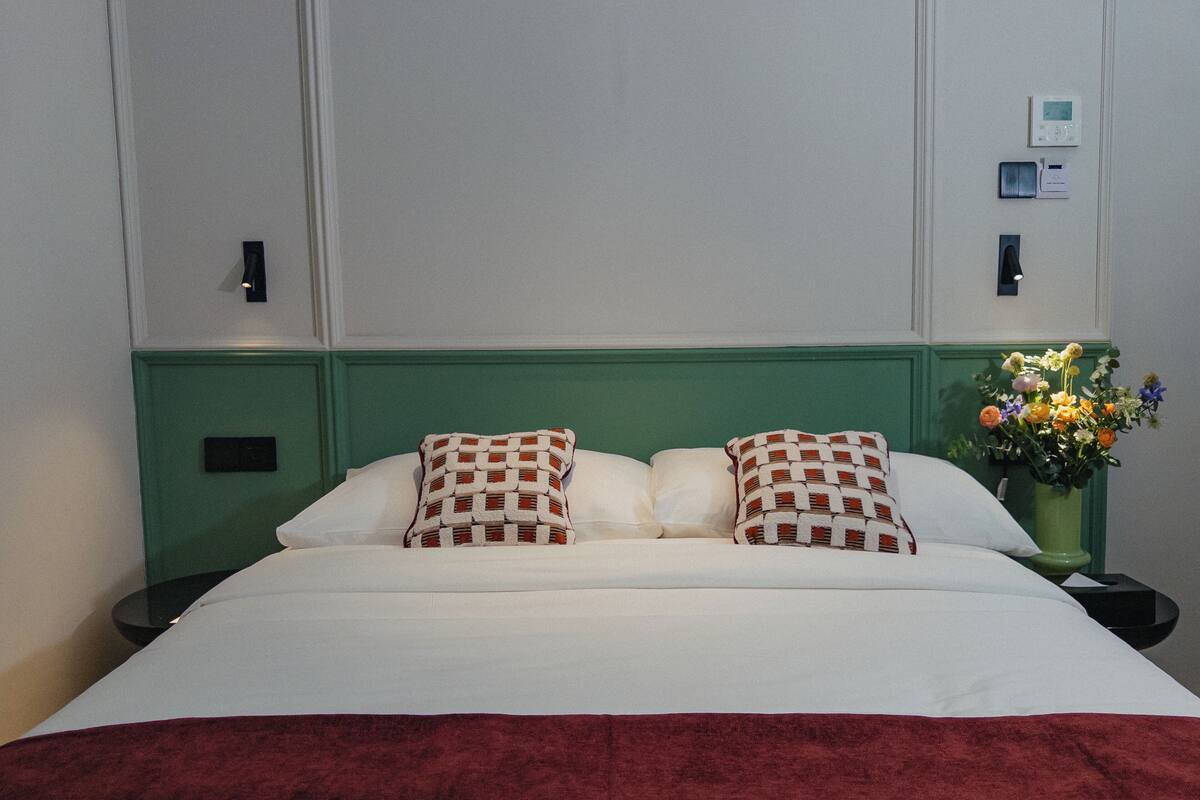 A neatly made bed with white linens and two patterned pillows, a green headboard, a nightstand with a lamp, and a vase with flowers.