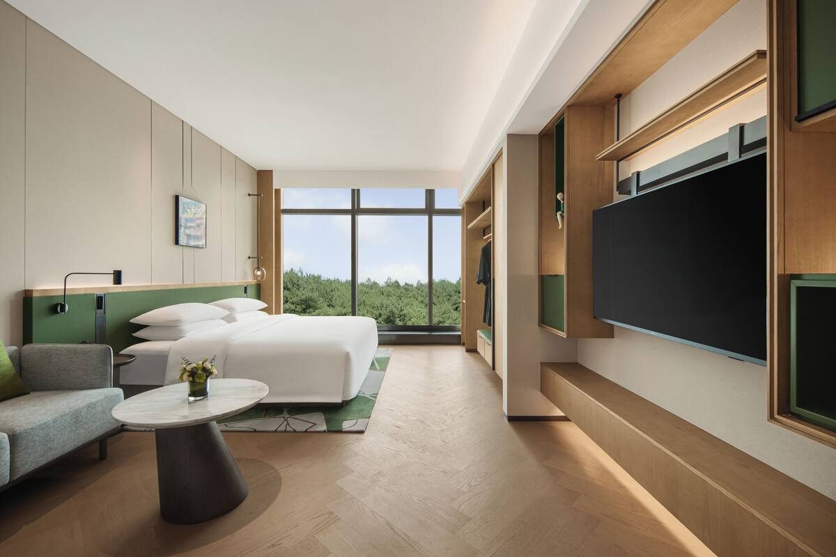 Habitación de hotel moderna con una cama grande, un sofá, una mesita redonda y un ventanal con vistas al exterior.