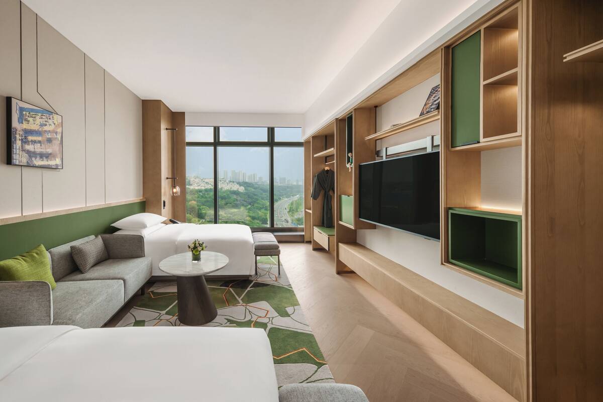 Una habitación de hotel moderna con una cama grande, un sofá, una mesita y un televisor. La habitación tiene un ventanal con vistas a la ciudad.