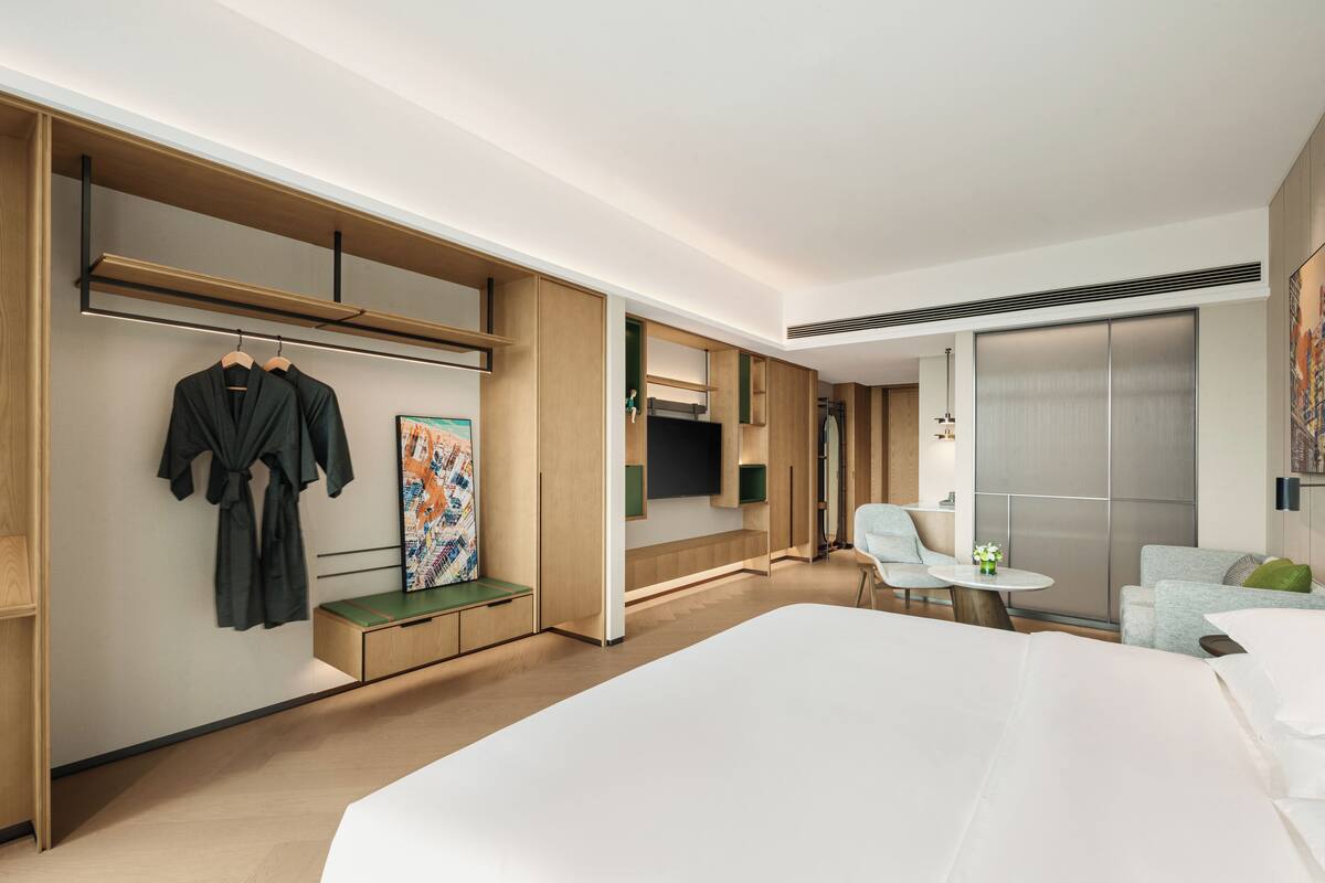 Una habitación de hotel moderna con una cama grande, un espacio de descanso con una mesita y sillas, un armario con ropa colgada y un espejo amplio.
