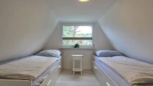 Ett litet, väl upplyst rum med två enkelsängar, ett litet vitt bord och ett fönster med utsikt över träd och hus.