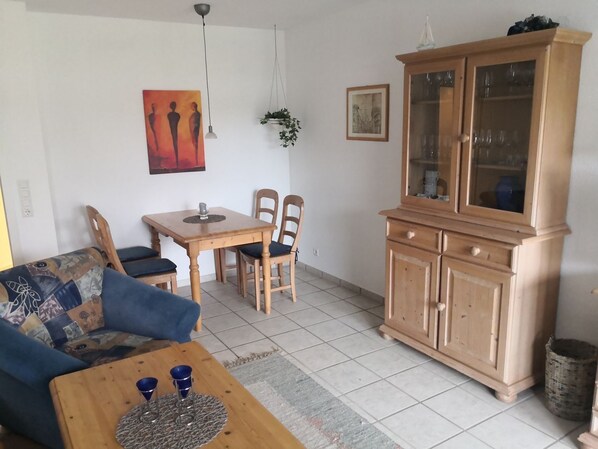 A living room with a sofa, a wooden coffee table, a dining table with chairs, a cabinet with glass doors, and artwork on the walls.