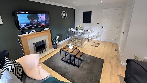 Un salon moderne avec une cheminée, un téléviseur à écran plat fixé au mur, un coin repas avec une table et des chaises en verre, et une porte blanche en arrière-plan.