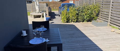 Una terraza de madera con una mesa preparada para dos, una silla de mimbre y una planta en maceta.