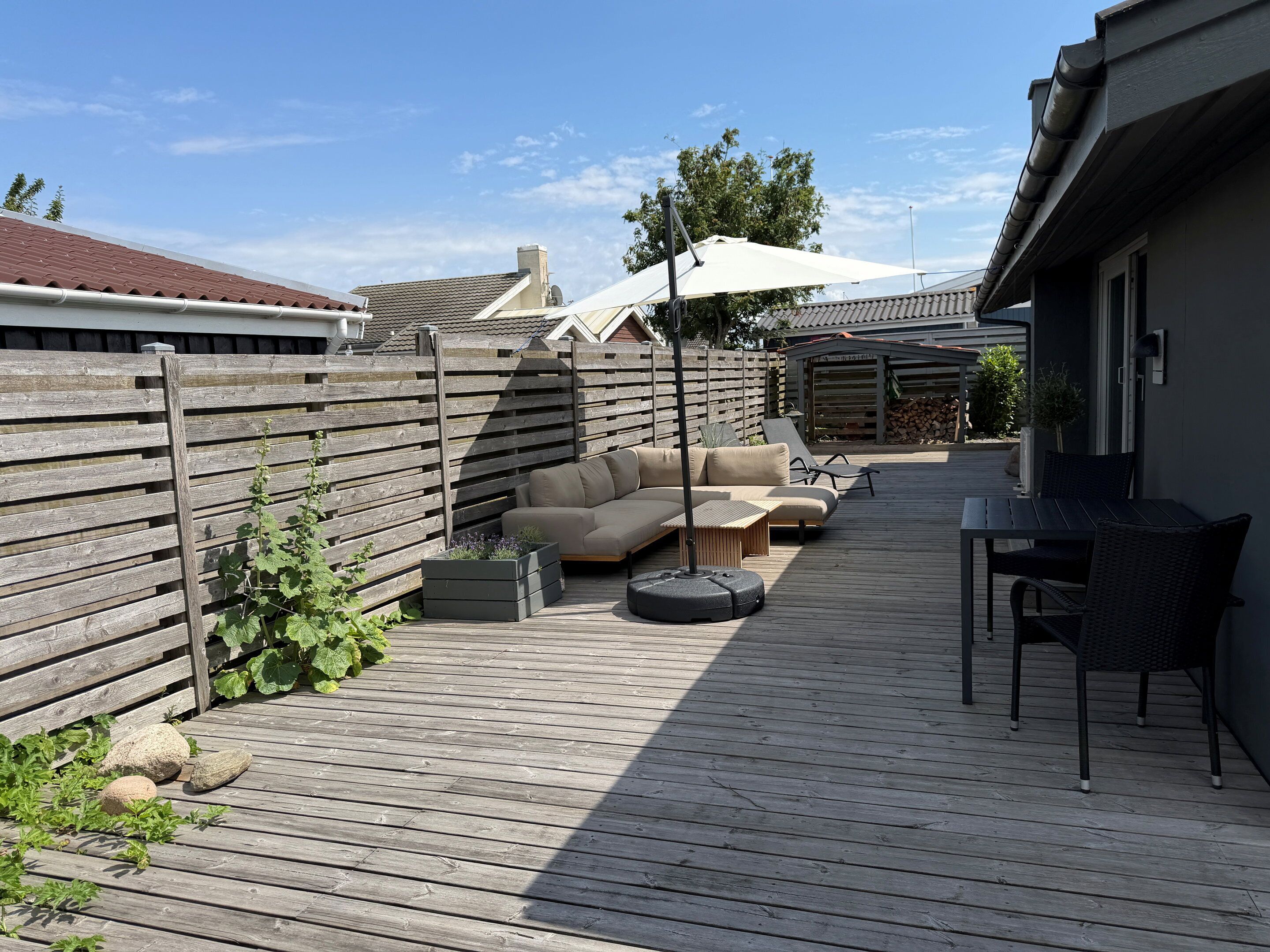 Eine Holzterrasse mit einer Couch und Stühlen, einem Tisch und einer Pergola mit Sonnenschirm.