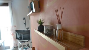 Una stanza con una TV su un mobile bianco, uno scaffale con libri e una pianta in vaso, e un diffusore con bastoncini di canna.