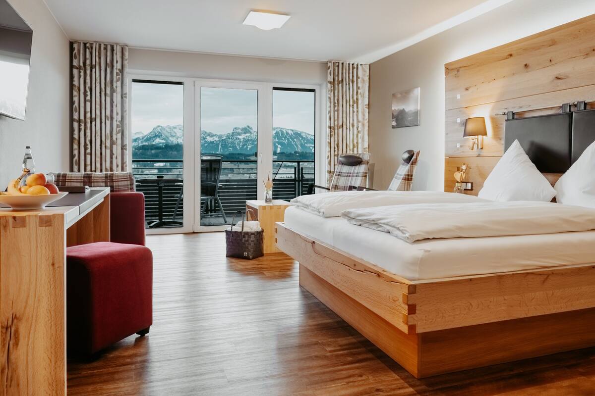 Een hotelkamer met een groot bed, een bureau met stoel, een kleine tafel met fruit, en uitzicht op bergen door de balkondeuren.