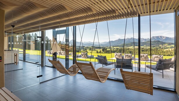 A modern interior with large windows, wooden ceiling slats, and outdoor seating by the glass walls. - Panorama Allgäu Spa Resort (Rueckholz)