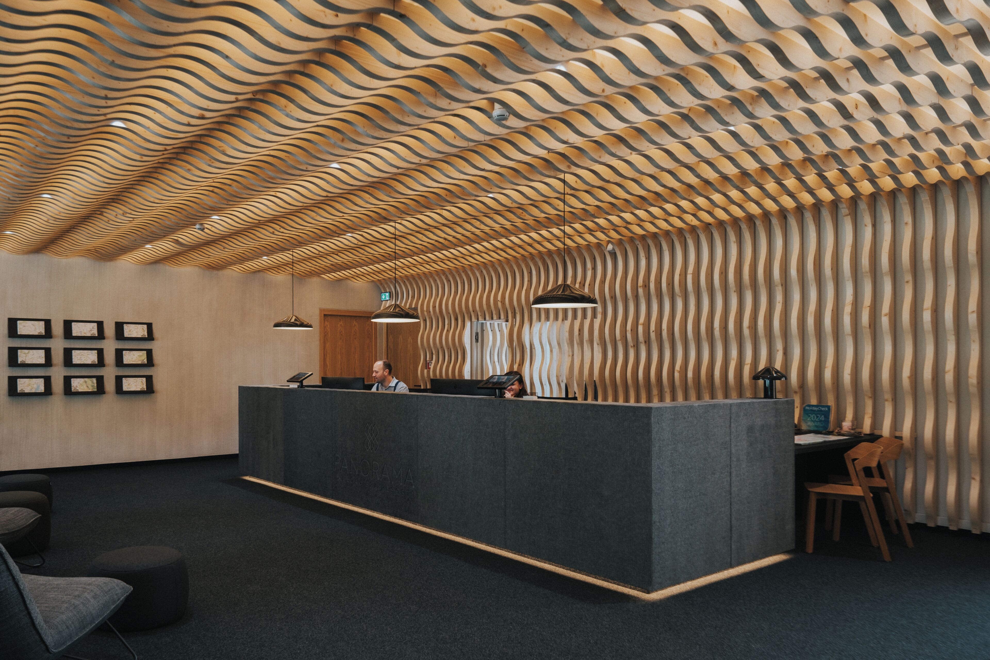 A reception area with a wavy wooden ceiling, a reception desk, framed pictures on the wall, and a seating area with a chair and ottoman.