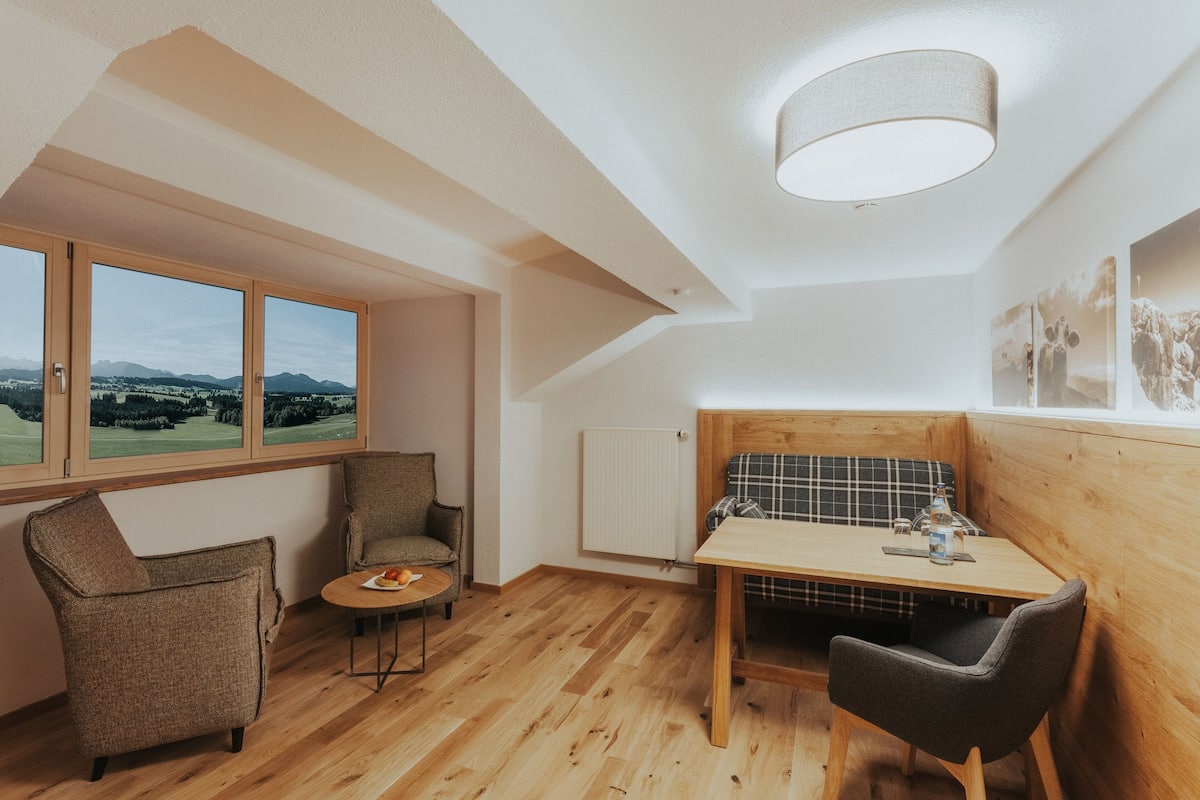 Een knus verblijf met een houten tafel, twee stoelen, uitzicht op bergen door grote ramen en een plafondlamp.