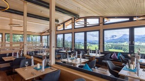 A restaurant with wooden interiors, large windows offering mountain views, and modern pendant lighting. - Panorama Allgäu Spa Resort (Rueckholz)