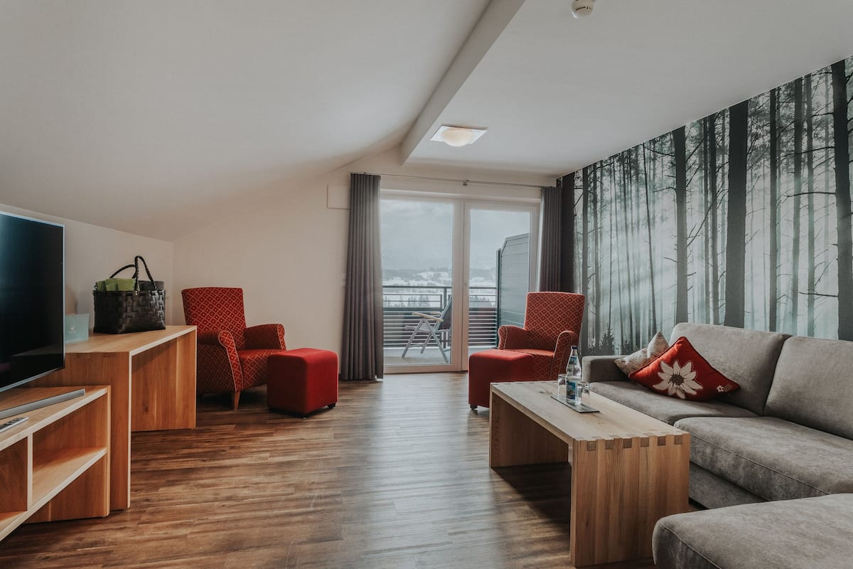 Een moderne woonkamer met houten vloer, rode en grijze meubels, een flatscreen televisie en een groot raam met uitzicht op een bos.
