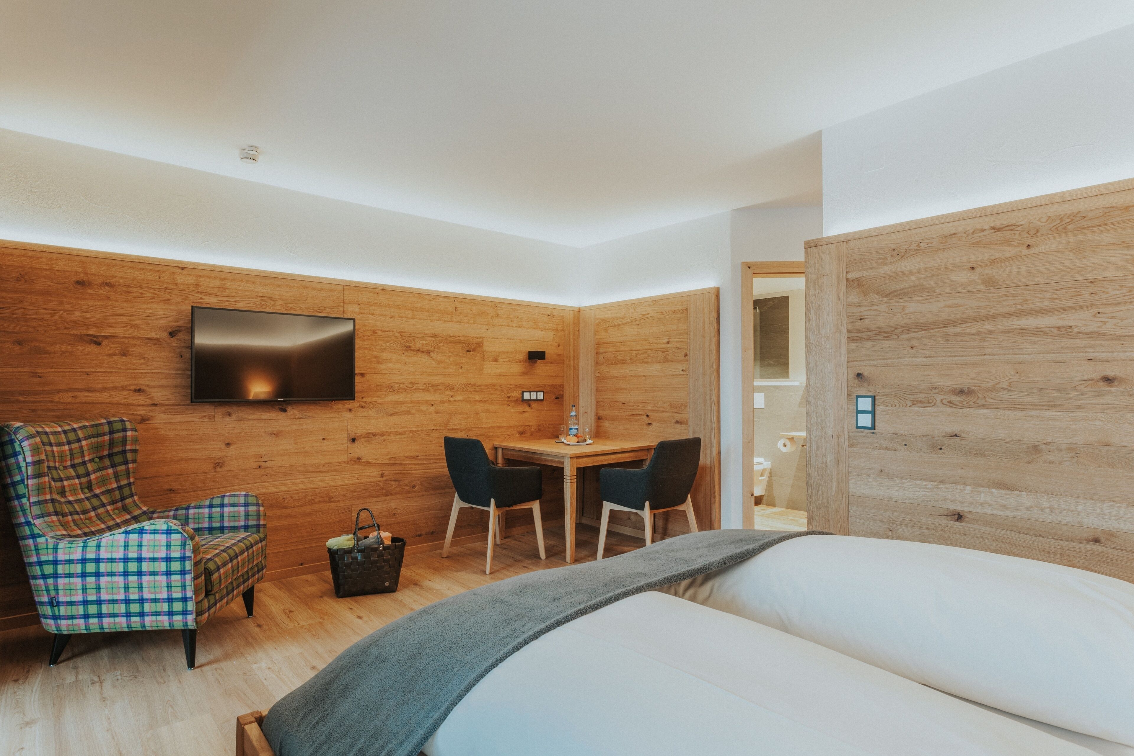 A modern room with a bed, a plaid armchair, a wooden desk with two chairs, and a flat-screen TV mounted on the wall.