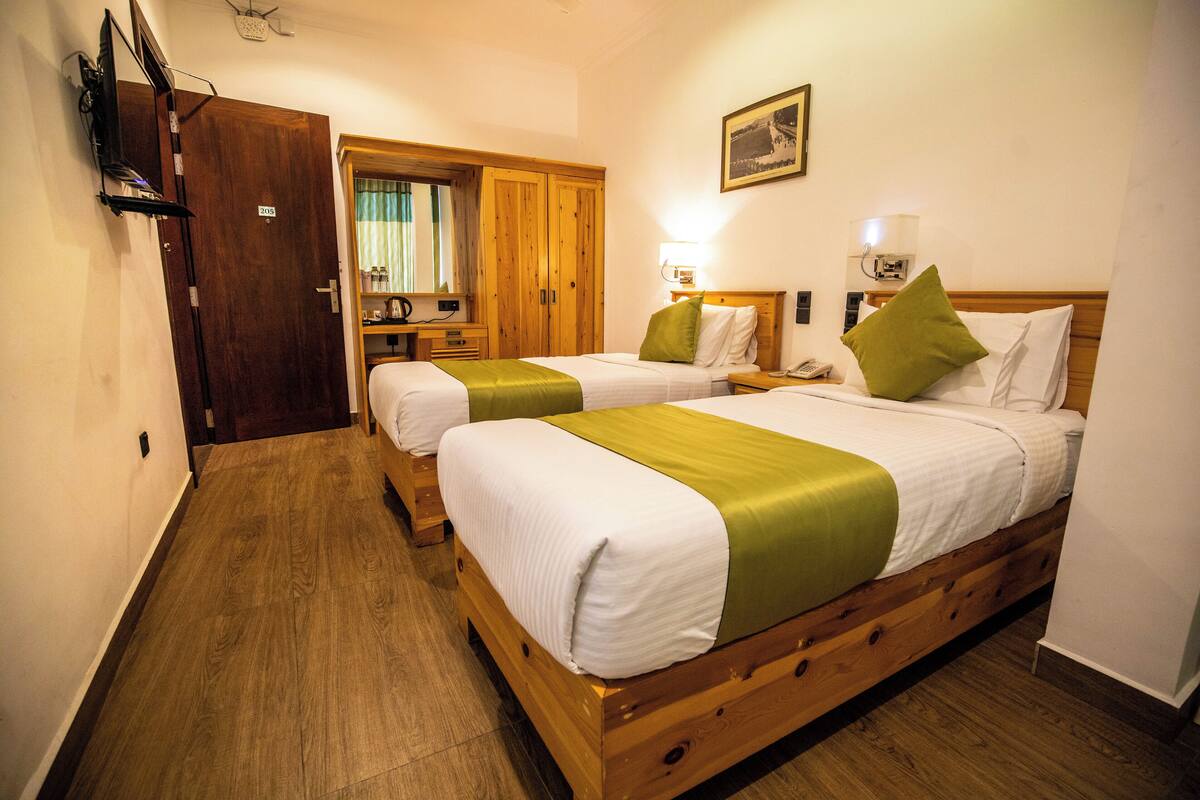 A hotel room with two beds, wooden flooring, a desk with a chair, a wardrobe, and a television on the wall.