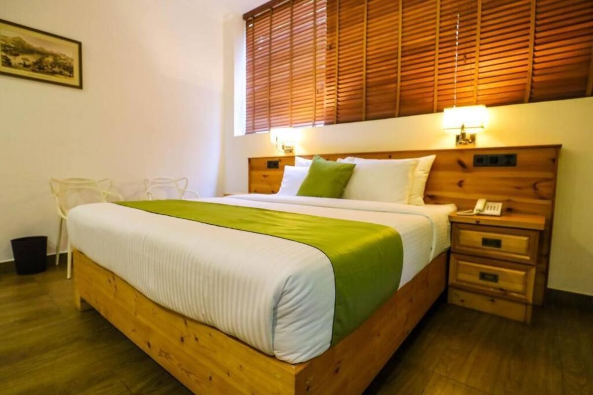 A wooden bed with a green and white bedspread, a bedside table with a lamp, and a window with wooden blinds.