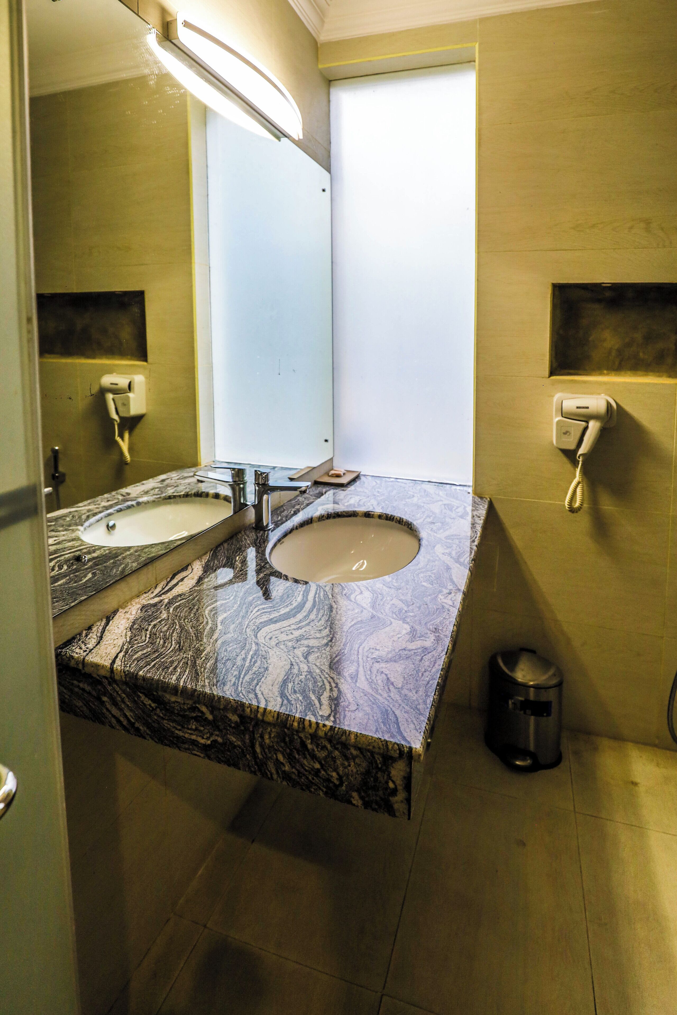 Un baño con lavamanos de mármol, un espejo, un secador de manos y un cubo de basura.