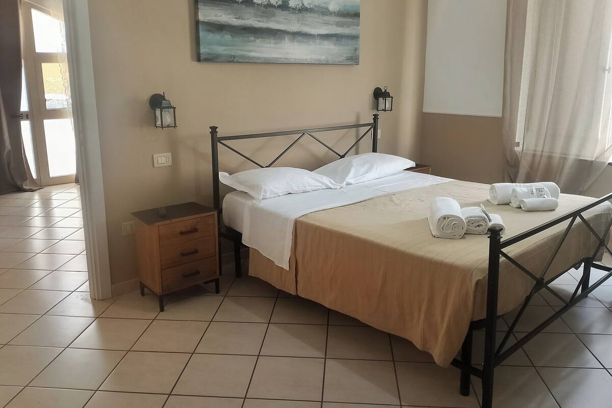 A neatly made bed with white linens, a bedside table with a lamp, and a painting on the wall.