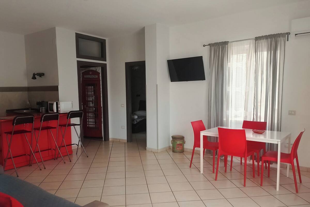 A compact living space with a kitchenette, a dining area with red chairs, a TV mounted on the wall, and a window with curtains.