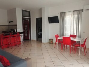 A compact living space with a kitchenette, a dining area with red chairs, a TV mounted on the wall, and a window with curtains. - Dedalo (Pompei)