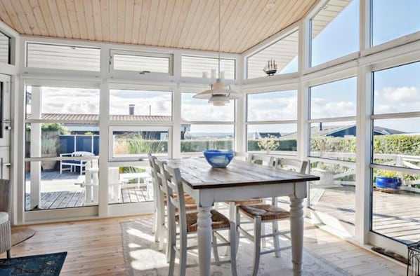 Une véranda avec une table et des chaises en bois, de grandes fenêtres et une vue sur l’extérieur.