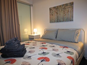 A neatly made bed with a floral bedspread, a stack of folded towels, a bedside lamp, and a painting on the wall. - 1BR Condo w/ Balcony/Pool/Gym (Toronto)