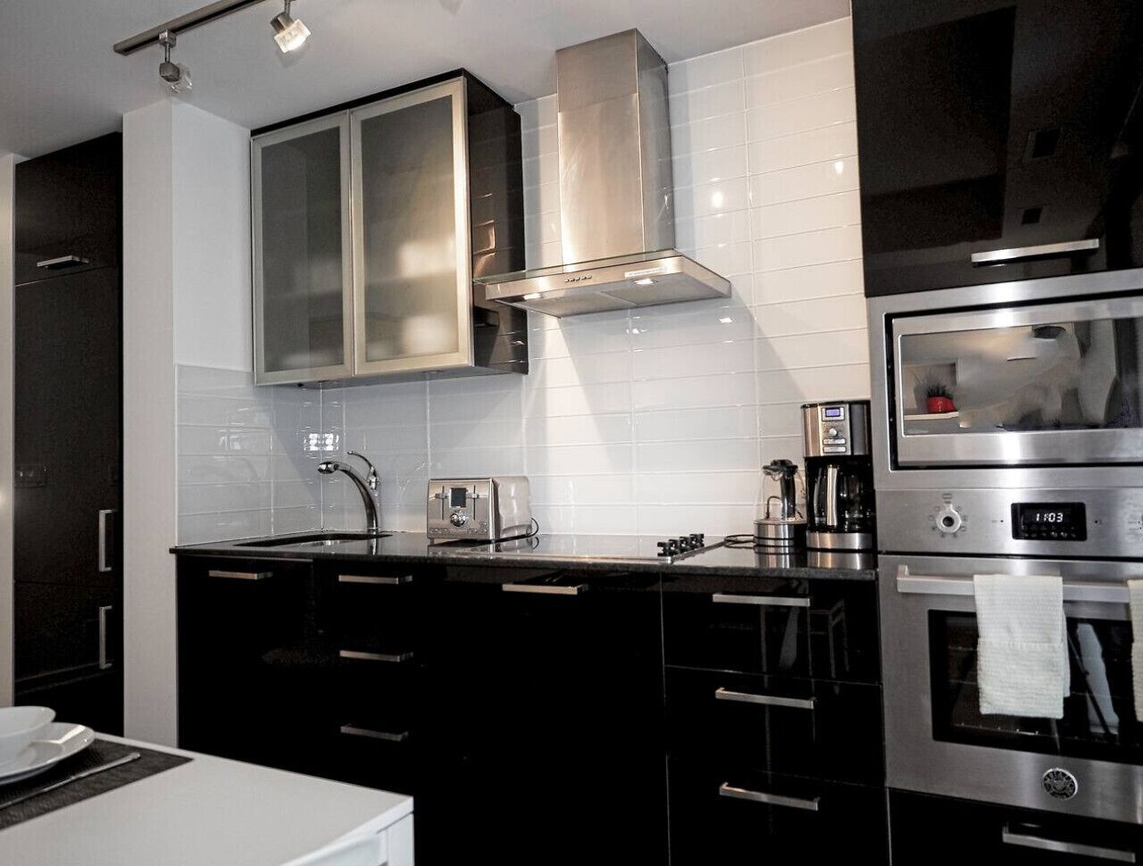 A modern kitchen with black cabinets, a stainless steel range hood, a built-in oven, and a coffee maker.